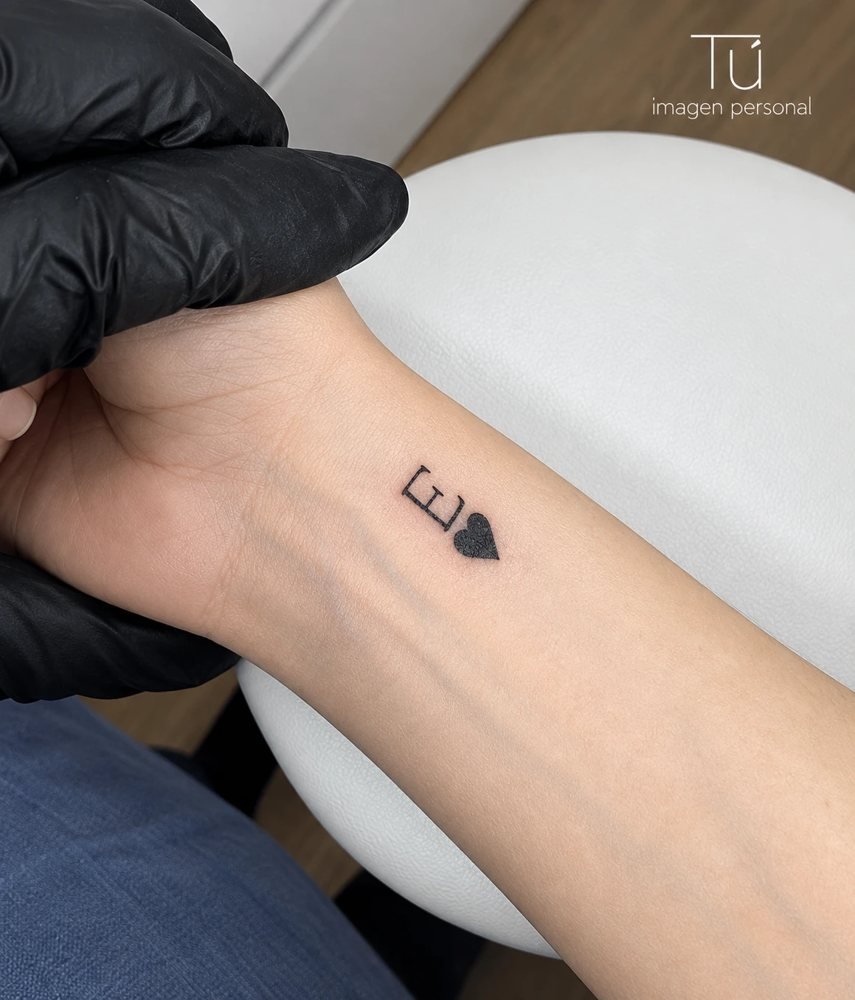 Tatuaje minimalista de la letra E con un corazón en la muñeca